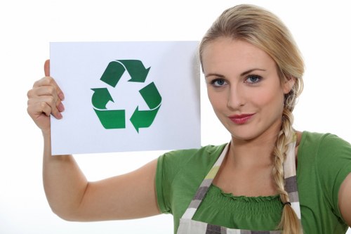 A young woman with blonde hair tied in a side braid is wearing a green top and a grey striped apron. She is standing outdoors against a plain white background and holding up a white rectangular sign with a large green recycling symbol composed of three chasing arrows. The woman’s expression is neutral, with a slight smile, and she is looking directly at the camera. The sign is positioned in front of her chest, with her right hand gripping the left side of the sign and her left hand supporting the right. The image emphasizes the recycling message, which aligns with environmentally conscious rubbish management practices, potentially highlighting the importance of recycling waste materials. The scene appears well-lit and clear, suitable for illustrations promoting responsible waste disposal and recycling efforts, possibly related to local waste management services like Commercial Waste Yeading in the area near Yeading, London, with a postcode around UB5 or UB4.