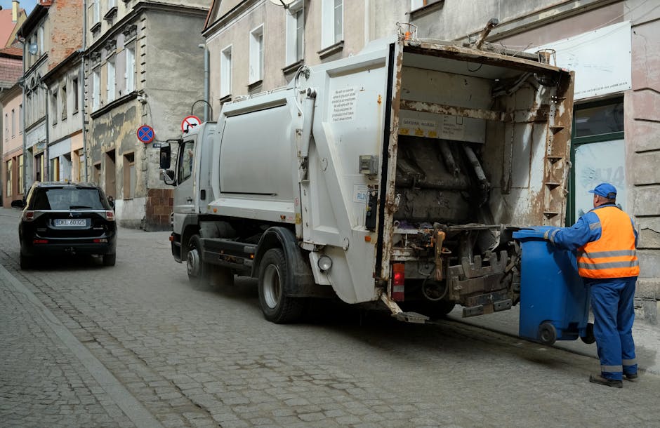 Five large black wheeled rubbish bins with yellow lids are lined up on a grey pavement against a plain light grey wall. Each bin has a white oval sticker with barcode and identifying numbers. The bins