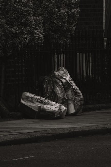 A pile of black plastic rubbish bags and discarded paper and cardboard debris stacked outside a red metal door on a concrete wall. The bags appear to contain household waste and are placed on a paveme
