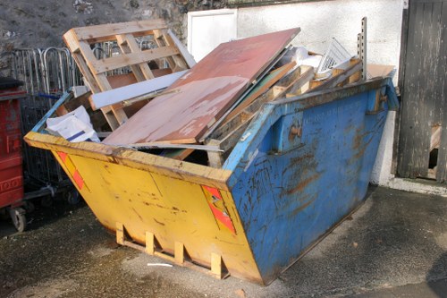 A large, rectangular metal skip painted in yellow with visible rust and scratches sits on a paved surface outdoors, filled with various discarded wooden pallets, boards, and panels, including a prominent brown wooden plank leaning against the top edge. The skip contains broken pieces of wood, some painted or stained, and miscellaneous construction debris, indicating unsorted waste awaiting removal. Behind the skip, a white wall provides a neutral backdrop, and to the right, part of a weathered wooden gate is visible. The environment appears to be a commercial or industrial area in Yeading, with the waste likely accumulated from renovation or manufacturing activities, reflecting the type of rubbish handled by Commercial Waste Yeading for efficient disposal or recycling services.