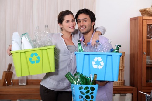 A young couple standing indoors in a domestic setting, holding three colour-coded recycling bins filled with glass bottles and recyclable waste. The woman on the left is holding a bright green bin with a recycling symbol, containing clear glass bottles, while the man on the right holds a blue bin with a recycling symbol, also filled with glass bottles. A third smaller blue bin, with a circular design, is in front of them and contains additional green glass bottles. They are standing in front of a wooden sideboard and a light-colored wall, with household items visible in the background. The overall environment suggests they are preparing for rubbish removal or recycling collection at a residence, consistent with waste management services in the Yeading area, near Postcode UB4, provided by Commercial Waste Yeading.