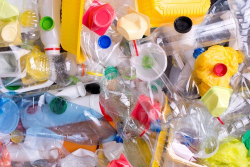 A large pile of discarded plastic bottles and containers, including clear, yellow, green, and red caps, with some bottles crushed or deformed, resting on a surface outdoors. The bottles vary in size and shape, with some having visible labels or markings. The collection includes empty soft drink bottles, water bottles, and other beverage containers, some with residual liquid or film inside. The background shows a mixture of different plastic waste items, with no visible packaging or other materials. The plastics appear used and discarded, indicative of waste collected for removal services such as those provided by Commercial Waste Yeading in the local town area. The overall scene emphasizes the need for rubbish collection and proper waste management, with the waste occupying a noticeable portion of an outdoor or possibly driveway setting, under bright natural light.